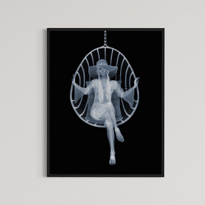 Framed Chromaluxe black and white x-ray photograph by Nick Veasey of a skeleton woman sitting in a swing chair with long pigtails, large hat and boho style dress crossing her legs