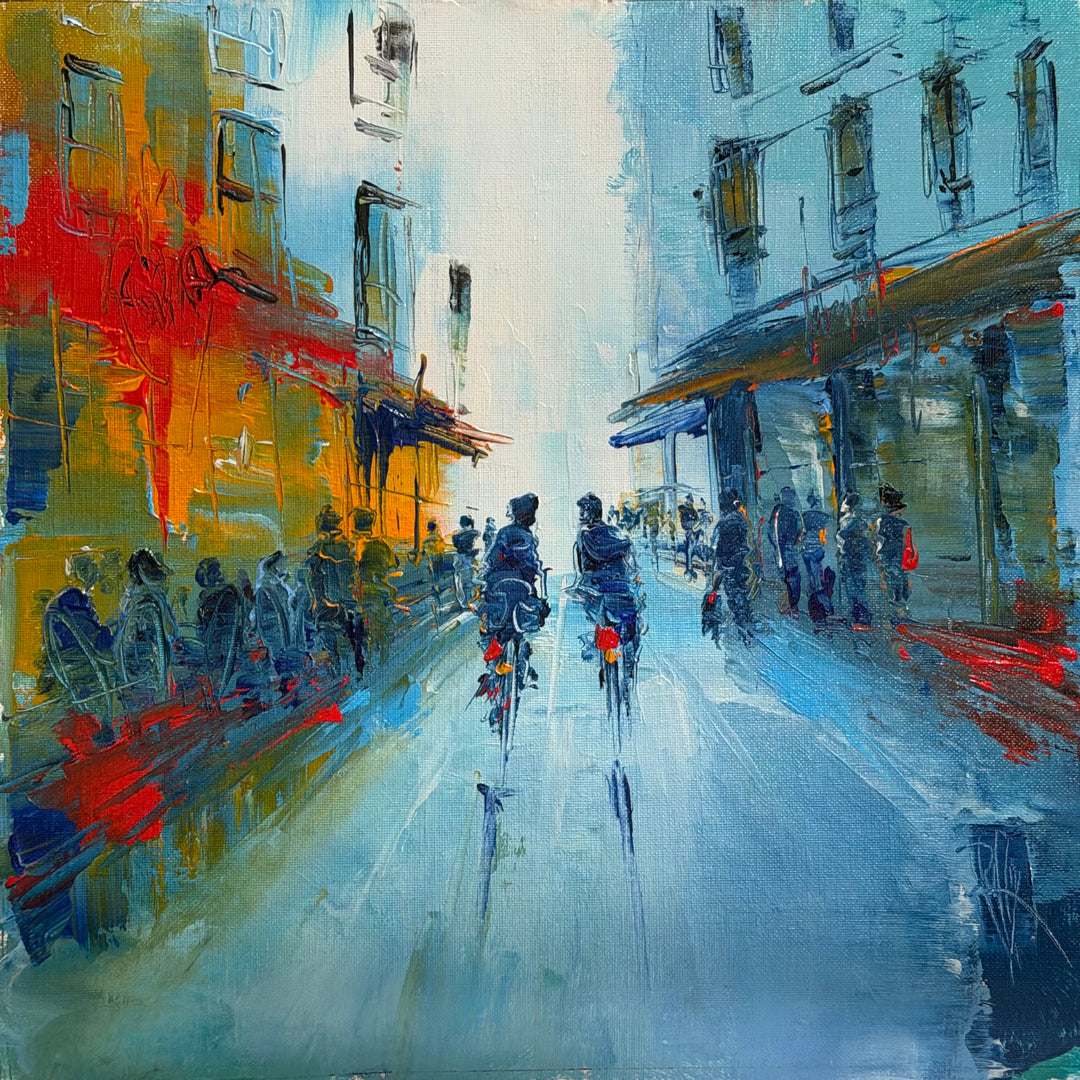 Christian Raffin, En Soirée, oil on canvas, 14 x 14 inches. An evening street with blue and orange buildings and lit shopfronts, small figures along both sides, and two cyclists riding down the center of a blue road.