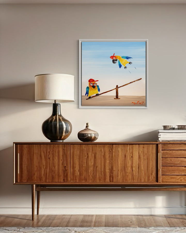 30” x 30” painting by French artist Moogly of two blue and yellow parrots with red baseball caps on a see-saw. One parrot is jumping on one end of the see-saw, making the other parrot fly up in the air. The parrots are made out of resin, giving them a textured and shiny finish. Their eyes are made out of washers that the artist has adhered to the canvas.